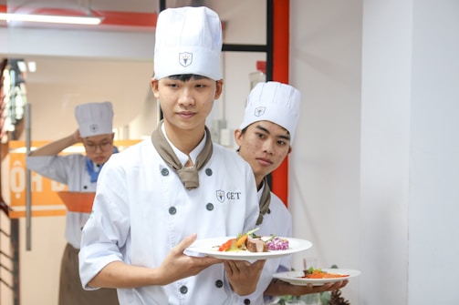 Three young chefs in white uniforms and hats are holding plates of beautifully arranged food. They appear to be in a kitchen or culinary setting, possibly near a cooking class or workshop area.