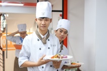 Three young chefs in white uniforms and hats are holding plates of beautifully arranged food. They appear to be in a kitchen or culinary setting, possibly near a cooking class or workshop area.