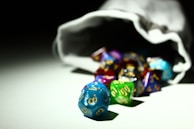 A collection of colorful, multi-sided dice spilling out of a white drawstring bag. The dice are in various colors including blue, green, purple, and red with gold numbers on them, and they rest on a white surface under a focused light source that creates contrasting shadows.