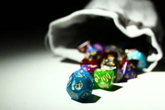 A collection of colorful, multi-sided dice spilling out of a white drawstring bag. The dice are in various colors including blue, green, purple, and red with gold numbers on them, and they rest on a white surface under a focused light source that creates contrasting shadows.