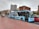 A city street scene featuring a blue and white AeroBus parked at a bus stop. The bus has signage for Barcelona El Prat Airport and offers free WiFi. Nearby, there are parked cars and a promotional poster in a glass-covered bus shelter. The buildings in the background are primarily brick-colored with various architectural styles. The street and sidewalk surface have a textured, patterned design.