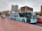 A city street scene featuring a blue and white AeroBus parked at a bus stop. The bus has signage for Barcelona El Prat Airport and offers free WiFi. Nearby, there are parked cars and a promotional poster in a glass-covered bus shelter. The buildings in the background are primarily brick-colored with various architectural styles. The street and sidewalk surface have a textured, patterned design.