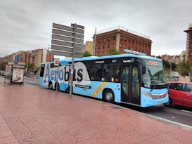 A city street scene featuring a blue and white AeroBus parked at a bus stop. The bus has signage for Barcelona El Prat Airport and offers free WiFi. Nearby, there are parked cars and a promotional poster in a glass-covered bus shelter. The buildings in the background are primarily brick-colored with various architectural styles. The street and sidewalk surface have a textured, patterned design.
