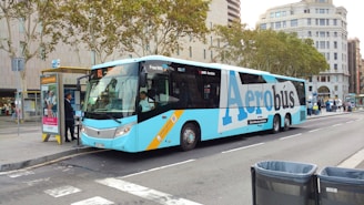 A blue and white Aerobus is parked at a bus stop on a city street. The bus features large branding along its side and offers 'Free WiFi' as indicated by a sign on the windshield. There is an advertisement at the bus stop and two individuals standing nearby, possibly passengers or bystanders. Trees with autumn leaves and various urban buildings surround the area, creating a typical cityscape.