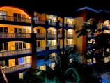 A multi-story hotel building illuminated at night with well-lit balconies and windows. Palm trees are visible in the foreground, casting silhouettes against the building.