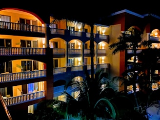 A multi-story hotel building illuminated at night with well-lit balconies and windows. Palm trees are visible in the foreground, casting silhouettes against the building.