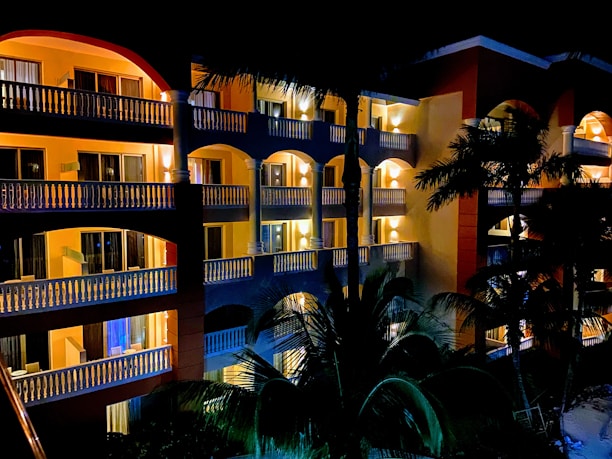 A multi-story hotel building illuminated at night with well-lit balconies and windows. Palm trees are visible in the foreground, casting silhouettes against the building.