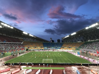 An expansive sports stadium with a soccer match taking place on a green, well-lit field. The stands are filled with spectators, some areas more densely populated than others. The structure is modern with a high roof and colorful seating arrangements, including red, yellow, and green chairs. The sky is a dramatic blend of blues and purples with scattered clouds, suggesting evening time.