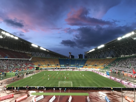 An expansive sports stadium with a soccer match taking place on a green, well-lit field. The stands are filled with spectators, some areas more densely populated than others. The structure is modern with a high roof and colorful seating arrangements, including red, yellow, and green chairs. The sky is a dramatic blend of blues and purples with scattered clouds, suggesting evening time.