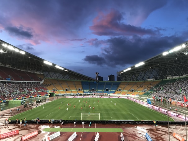 An expansive sports stadium with a soccer match taking place on a green, well-lit field. The stands are filled with spectators, some areas more densely populated than others. The structure is modern with a high roof and colorful seating arrangements, including red, yellow, and green chairs. The sky is a dramatic blend of blues and purples with scattered clouds, suggesting evening time.