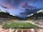 An expansive sports stadium with a soccer match taking place on a green, well-lit field. The stands are filled with spectators, some areas more densely populated than others. The structure is modern with a high roof and colorful seating arrangements, including red, yellow, and green chairs. The sky is a dramatic blend of blues and purples with scattered clouds, suggesting evening time.