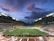 An expansive sports stadium with a soccer match taking place on a green, well-lit field. The stands are filled with spectators, some areas more densely populated than others. The structure is modern with a high roof and colorful seating arrangements, including red, yellow, and green chairs. The sky is a dramatic blend of blues and purples with scattered clouds, suggesting evening time.