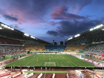 An expansive sports stadium with a soccer match taking place on a green, well-lit field. The stands are filled with spectators, some areas more densely populated than others. The structure is modern with a high roof and colorful seating arrangements, including red, yellow, and green chairs. The sky is a dramatic blend of blues and purples with scattered clouds, suggesting evening time.