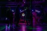 Silhouettes of people dancing in a dimly lit room with colorful, neon lighting. A neon sign displays '50's Dance' in the background, and there are balloons in various shades of pink and white on one side. The scene captures a lively and retro-themed dance event.