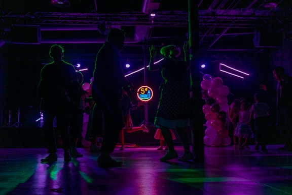 Silhouettes of people dancing in a dimly lit room with colorful, neon lighting. A neon sign displays '50's Dance' in the background, and there are balloons in various shades of pink and white on one side. The scene captures a lively and retro-themed dance event.