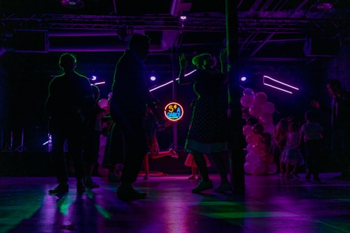 Silhouettes of people dancing in a dimly lit room with colorful, neon lighting. A neon sign displays '50's Dance' in the background, and there are balloons in various shades of pink and white on one side. The scene captures a lively and retro-themed dance event.