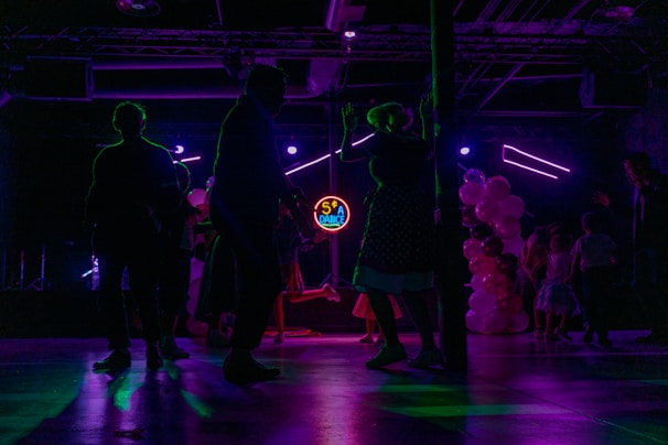 Silhouettes of people dancing in a dimly lit room with colorful, neon lighting. A neon sign displays '50's Dance' in the background, and there are balloons in various shades of pink and white on one side. The scene captures a lively and retro-themed dance event.