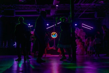 Silhouettes of people dancing in a dimly lit room with colorful, neon lighting. A neon sign displays '50's Dance' in the background, and there are balloons in various shades of pink and white on one side. The scene captures a lively and retro-themed dance event.