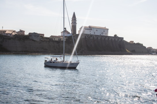 A sailboat glides across a shimmering body of water against the backdrop of a historic coastal town. The warm sunlight casts a soft glow on the buildings perched on a bluff, with a prominent church tower rising above them. The sea sparkles under the clear sky, creating a serene and picturesque scene.