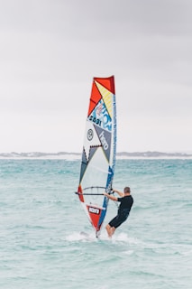 A person windsurfing on clear turquoise water with a colorful sail featuring geometric patterns and logos. The sky is overcast and the background shows a low shoreline.