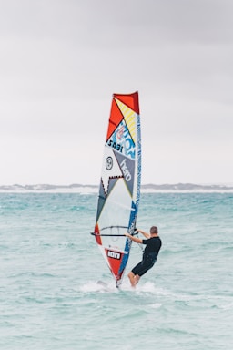 A person windsurfing on clear turquoise water with a colorful sail featuring geometric patterns and logos. The sky is overcast and the background shows a low shoreline.