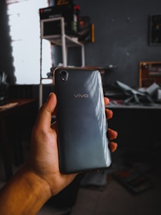 A hand holding a smartphone with the brand name Vivo visible. The phone has a black or dark gray back with slightly curved edges. The background shows a dimly lit room with shelves and scattered items, creating a casual and somewhat cluttered environment.