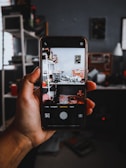 A hand holding a smartphone showing a camera app interface capturing a cluttered room with shelves and various items. The background features a dark room with shelves and objects.