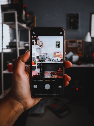 A hand holding a smartphone showing a camera app interface capturing a cluttered room with shelves and various items. The background features a dark room with shelves and objects.