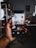 A hand holding a smartphone showing a camera app interface capturing a cluttered room with shelves and various items. The background features a dark room with shelves and objects.
