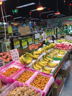 A bustling fruit section in a grocery store with an array of fresh produce neatly arranged. There are bananas, peaches, and other tropical fruits displayed in colorful baskets. Various price tags are seen in front of the fruits, and overhead red lamps illuminate the area. The atmosphere appears lively with several people browsing around.