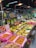 A bustling fruit section in a grocery store with an array of fresh produce neatly arranged. There are bananas, peaches, and other tropical fruits displayed in colorful baskets. Various price tags are seen in front of the fruits, and overhead red lamps illuminate the area. The atmosphere appears lively with several people browsing around.