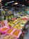 A bustling fruit section in a grocery store with an array of fresh produce neatly arranged. There are bananas, peaches, and other tropical fruits displayed in colorful baskets. Various price tags are seen in front of the fruits, and overhead red lamps illuminate the area. The atmosphere appears lively with several people browsing around.