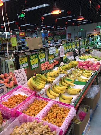 A bustling fruit section in a grocery store with an array of fresh produce neatly arranged. There are bananas, peaches, and other tropical fruits displayed in colorful baskets. Various price tags are seen in front of the fruits, and overhead red lamps illuminate the area. The atmosphere appears lively with several people browsing around.