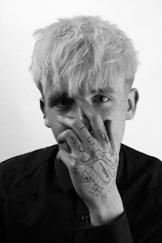 A person with light-colored hair is covering part of their face with their hand. The hand has various black ink tattoos, including abstract shapes and symbols. The individual is wearing a dark shirt, and the overall photography is in black and white.
