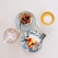 A selection of desserts and drinks arranged on a light surface, including a slice of fruit-topped cake on a blue and white floral plate, a small baked dessert with berries on a lattice-patterned plate, a cup of cappuccino with latte art, and an iced drink with a straw.