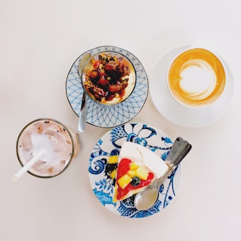 A selection of desserts and drinks arranged on a light surface, including a slice of fruit-topped cake on a blue and white floral plate, a small baked dessert with berries on a lattice-patterned plate, a cup of cappuccino with latte art, and an iced drink with a straw.