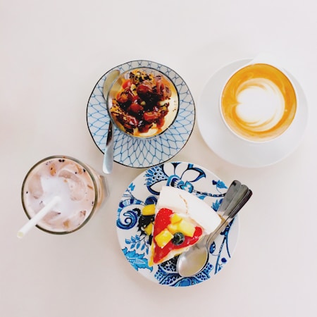 A selection of desserts and drinks arranged on a light surface, including a slice of fruit-topped cake on a blue and white floral plate, a small baked dessert with berries on a lattice-patterned plate, a cup of cappuccino with latte art, and an iced drink with a straw.