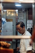 A man in a white chef's uniform is standing in a small, dimly lit kitchen space, focused on his work. Visible in the background are kitchen equipment, a calendar on the wall, and switches on a panel.