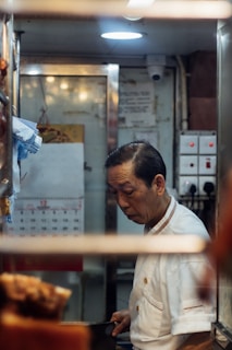 A man in a white chef's uniform is standing in a small, dimly lit kitchen space, focused on his work. Visible in the background are kitchen equipment, a calendar on the wall, and switches on a panel.