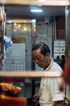 A man in a white chef's uniform is standing in a small, dimly lit kitchen space, focused on his work. Visible in the background are kitchen equipment, a calendar on the wall, and switches on a panel.