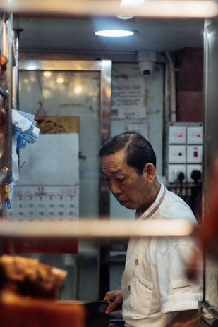 A man in a white chef's uniform is standing in a small, dimly lit kitchen space, focused on his work. Visible in the background are kitchen equipment, a calendar on the wall, and switches on a panel.