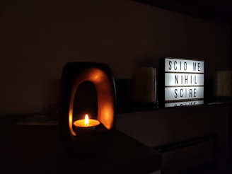 A candle glows softly inside a dark holder on a shelf, casting a warm, intimate light. Beside it, an illuminated lightbox displays the message 'SCIO ME NIHIL SCIRE', surrounded by dimly lit candles in the background.
