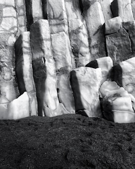 Rugged basalt columns rise vertically, exhibiting varied textures from smooth to rough. The surface is interrupted by cracks and crevices, casting shadows that emphasize the geometric structure. At the base, dark sand or gravel provides a contrasting foreground.