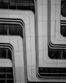 A modern architectural facade features sleek, overlapping panels with smooth, curved lines. The surface appears metallic and is dotted with small rivets, contributing to a futuristic look. Reflective glass sections are visible, partially revealing the building's interior.