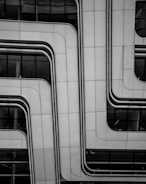 A modern architectural facade features sleek, overlapping panels with smooth, curved lines. The surface appears metallic and is dotted with small rivets, contributing to a futuristic look. Reflective glass sections are visible, partially revealing the building's interior.