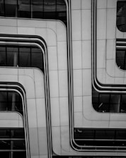 A modern architectural facade features sleek, overlapping panels with smooth, curved lines. The surface appears metallic and is dotted with small rivets, contributing to a futuristic look. Reflective glass sections are visible, partially revealing the building's interior.