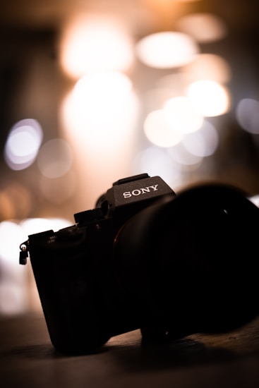 A camera with prominent branding is positioned at an angle, set against a backdrop of blurred lights, creating a bokeh effect.