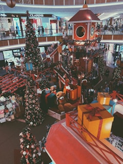 A festive Christmas display in a shopping mall featuring large gift boxes, decorated Christmas trees, and a Santa's workshop theme with a large clock tower and a giant teddy bear among the decorations. The scene includes elements of Christmas like lights, ornaments, and garlands, creating a bustling and lively atmosphere with people strolling along the upper and lower levels of the mall.