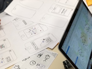 A series of drawn wireframes and sketches on white paper showcasing mobile app interface designs. The designs are accompanied by a laptop displaying a digital map with various data points. Pens and other sketching tools are partially visible, indicating a workspace focused on digital design and mapping.