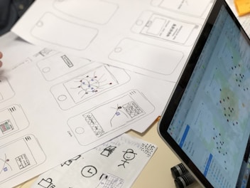 A series of drawn wireframes and sketches on white paper showcasing mobile app interface designs. The designs are accompanied by a laptop displaying a digital map with various data points. Pens and other sketching tools are partially visible, indicating a workspace focused on digital design and mapping.